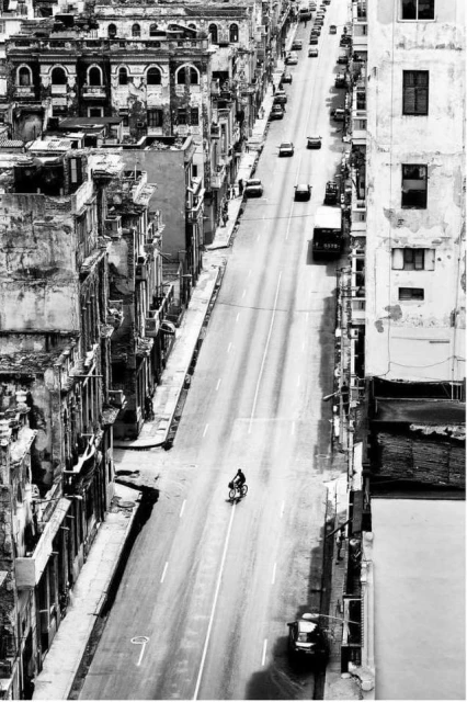 Black and white shot of a cyclist riding on a street lined with vintage cars and old buildings under sunlight in an urban setting.