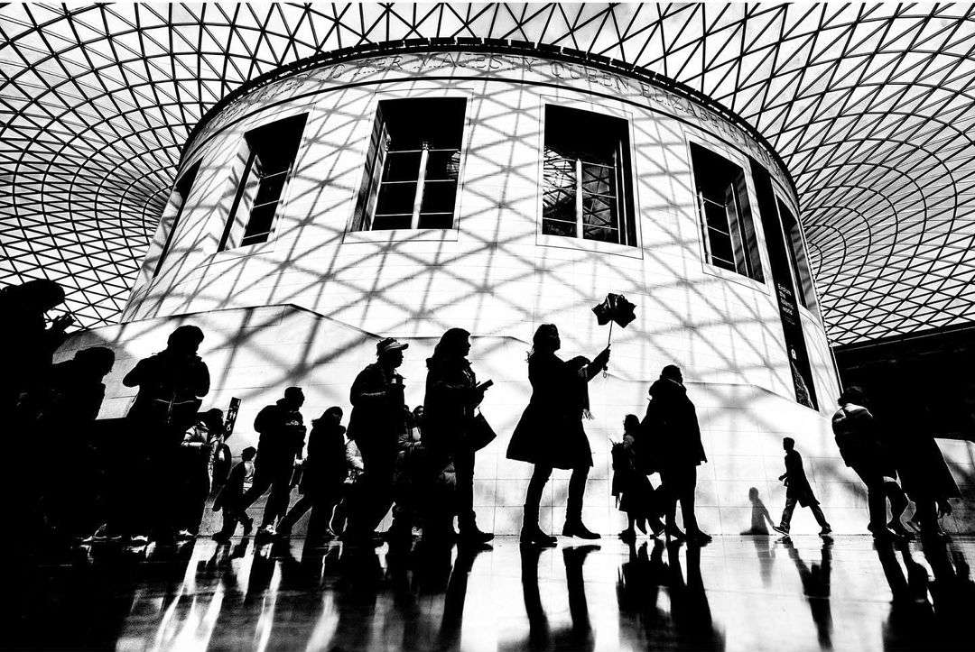 Black and white interior shot of a crowd of people walking under the geometric grid of the Queen Elizabeth Great Court roof in the British Museum.