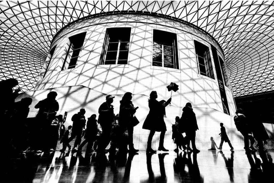 Black and white interior shot of a crowd of people walking under the geometric grid of the Queen Elizabeth Great Court roof in the British Museum.