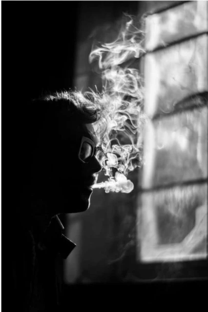 Monochrome portrait of a person exhaling smoke near a window, featuring dark tones and contrasting light creating a dramatic effect.