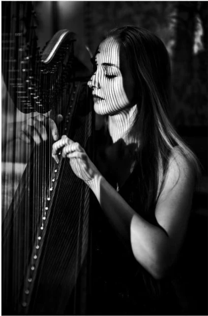 Black and white portrait of a woman playing a harp, with lines of light and shadow across her face, creating a dramatic and artistic effect.