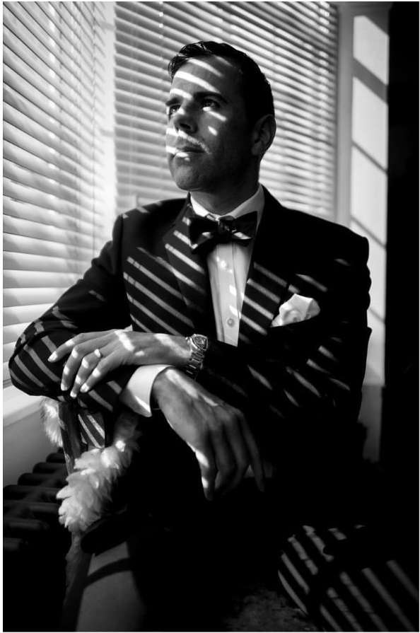 A man in a tuxedo sits near a window with light and shadow stripes, gazing to the side with a classic, elegant, and composed atmosphere.