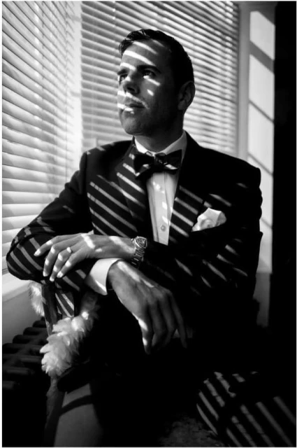 A man in a tuxedo sits near a window with light and shadow stripes, gazing to the side with a classic, elegant, and composed atmosphere.