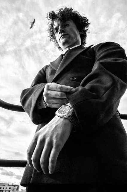Black and white portrait of a man with curly hair in a jacket, adjusting his cuff, with a bird and cloudy sky in the background.