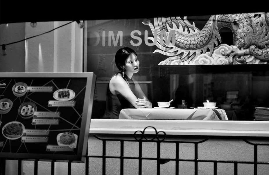 A woman is sitting at a table in front of a window with a dragon mural. A restaurant menu stands to the side in black and white.