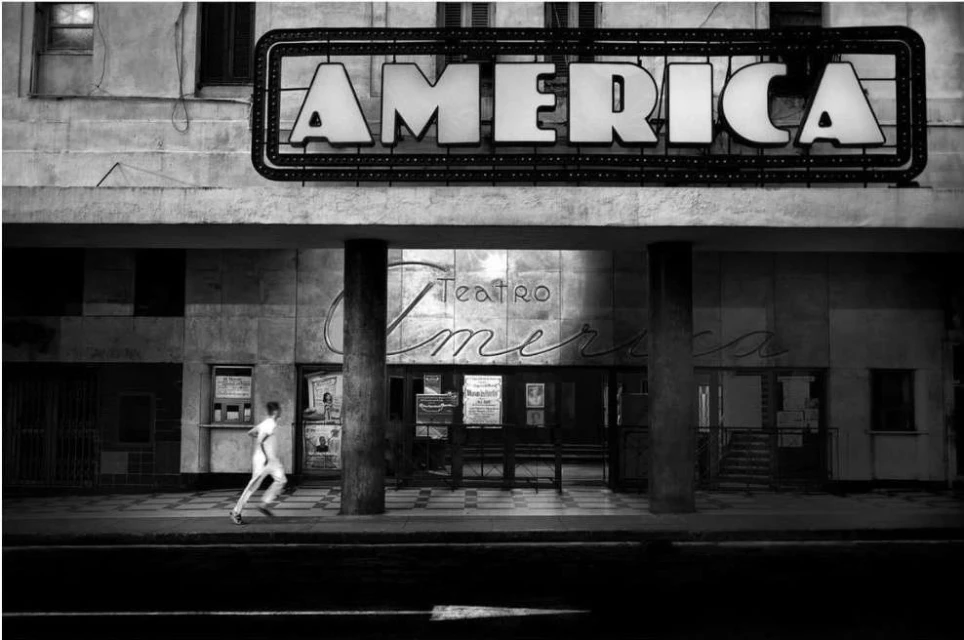 Black and white photograph of a vintage theater building with a man running past the front of the building on the sidewalk.