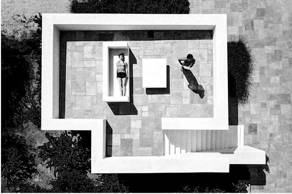 Aerial shot of an adult female standing near a woman reclining on a platform within a modern architectural space featuring tile flooring.