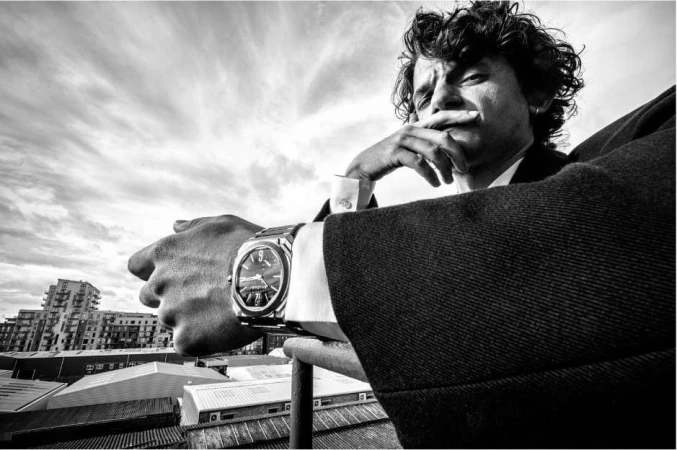 Monochrome portrait of a man leaning on a railing wearing a suit and a metallic wristwatch against an urban rooftop background.