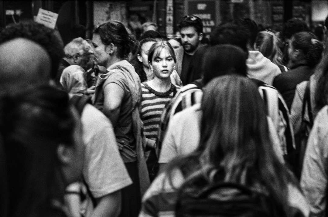 Black and white photo of a woman with striped shirt standing in a crowd, looking forward with a serious expression.