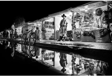 Monochrome street photography of a vendor standing outside lit market stalls with a bicycle and reflection in puddles