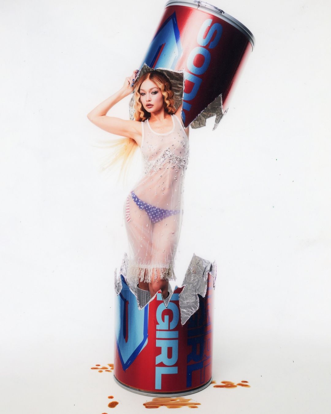 A blonde woman poses inside a shattered, oversized soda can, wearing a sheer, embellished dress against a clean, white backdrop.