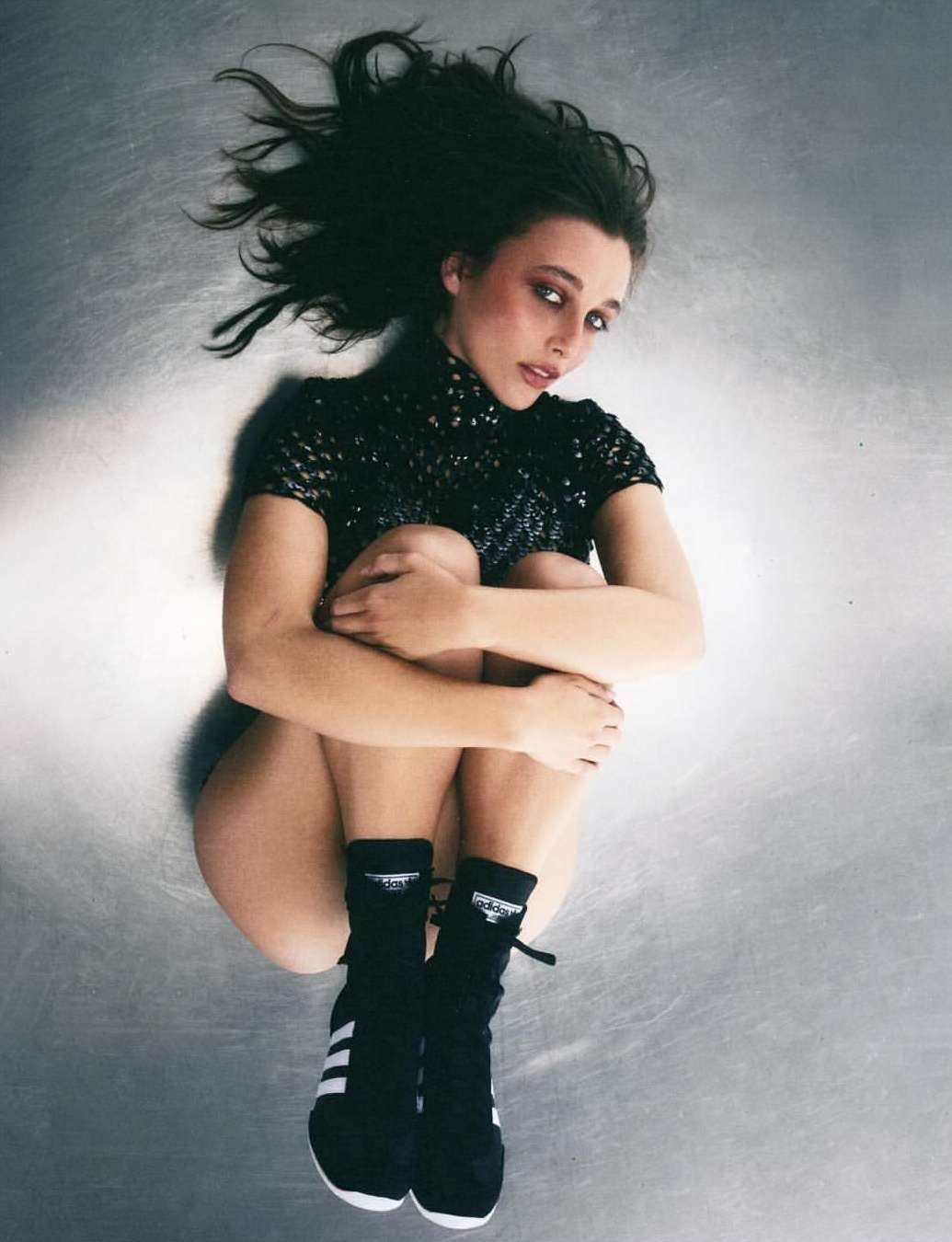 Young woman with dark hair and makeup hugs knees while laying on a mottled gray surface, wearing a black top and Adidas-branded socks and shoes.