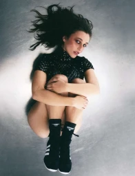 Woman wearing a black patterned top and black sneakers with white stripes, photographed from above against a textured metallic floor.