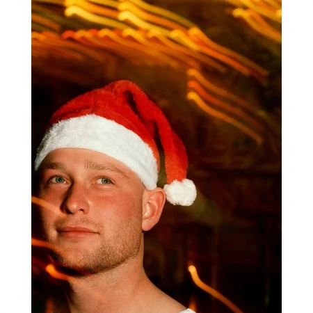 Portrait of a fair-skinned adult male wearing a red and white Santa Claus hat against a blurred orange background of holiday lights.