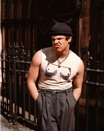 Young man in a black beanie with horns, graphic tank top, and pleated trousers standing outdoors near a wrought-iron fence.