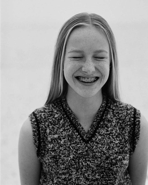Close-up black-and-white portrait of a smiling young woman with freckles and braces, eyes closed, wearing a textured sleeveless sweater.