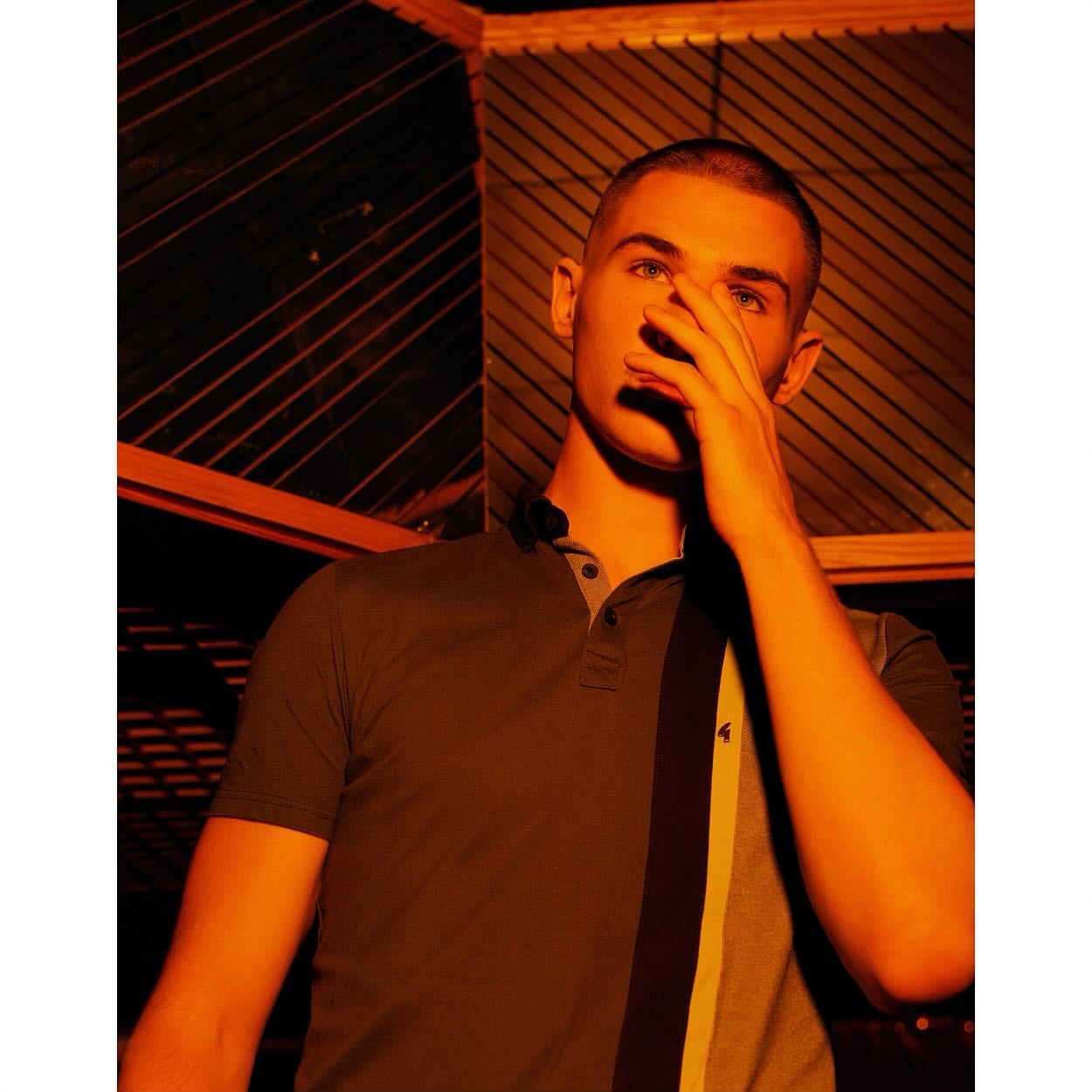 Portrait of a young adult male wearing a polo shirt, his face partially covered by his hand, indoor under an orange light source.