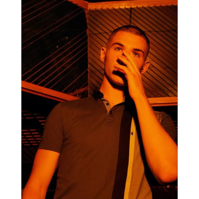 Portrait of a young adult male wearing a polo shirt, his face partially covered by his hand, indoor under an orange light source.
