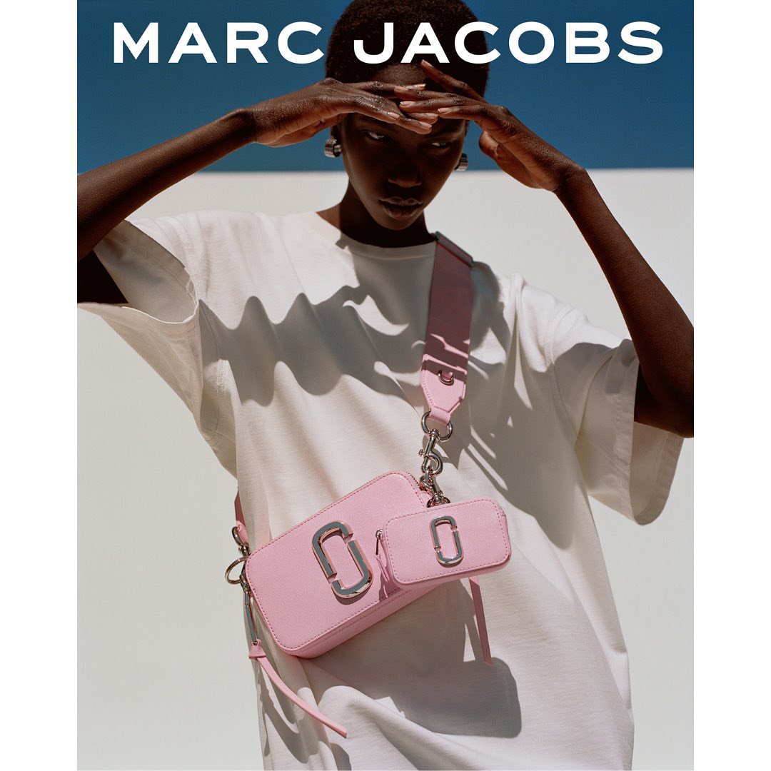 Model wearing a white t-shirt and pink crossbody bag poses against a white wall under a blue sky.