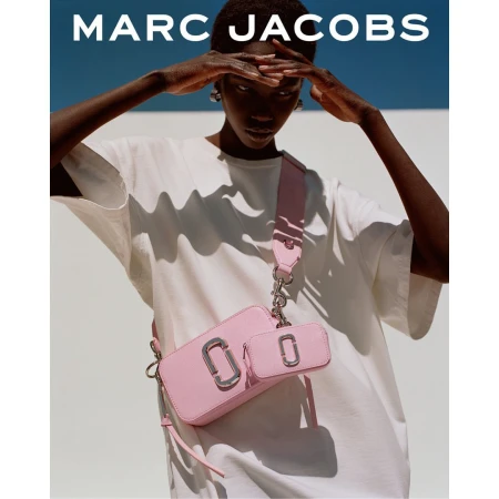 Model wearing a white t-shirt and pink crossbody bag poses against a white wall under a blue sky.