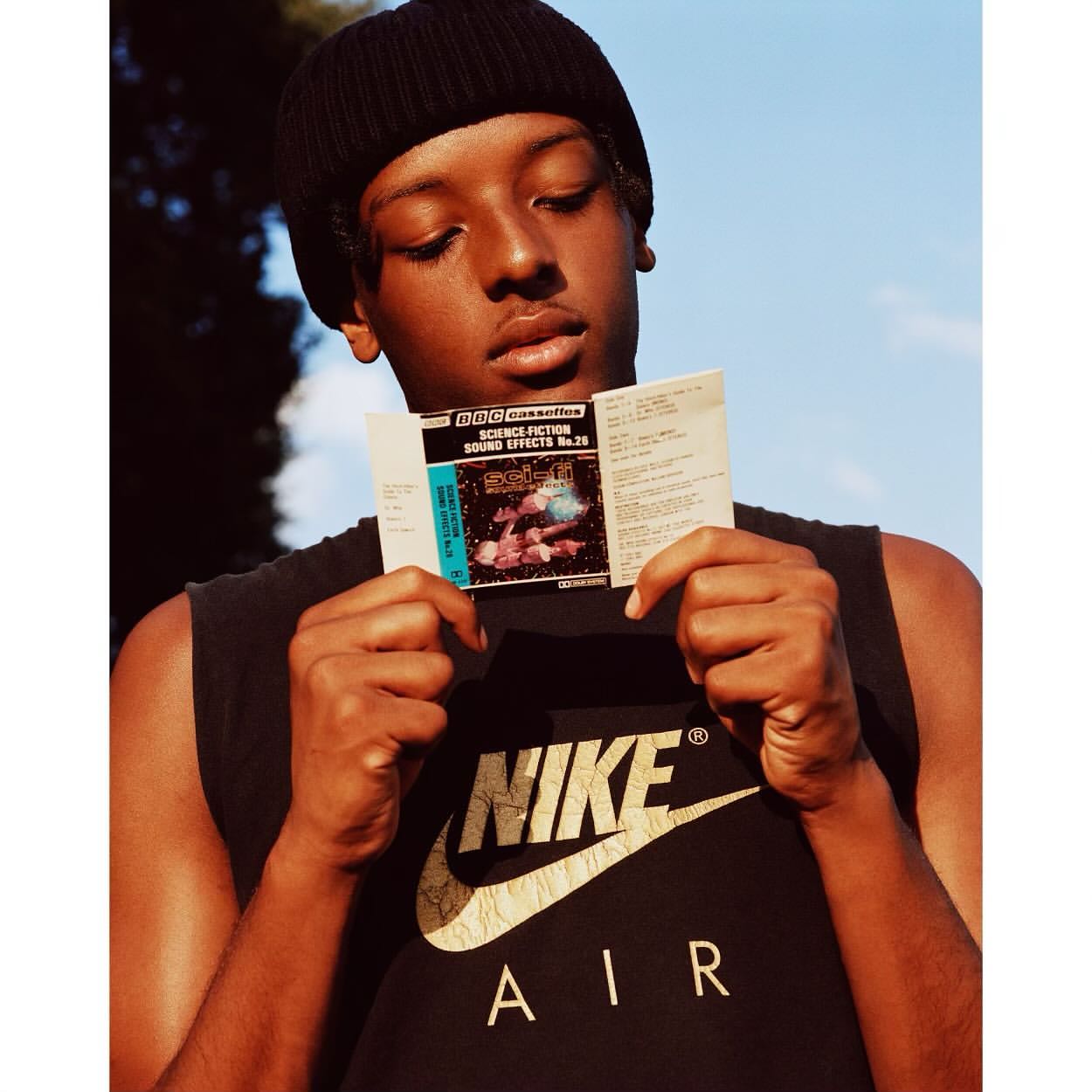 An adult with a black beanie holds a cassette tape case outdoors with trees and sky in the background, wearing a Nike t-shirt.
