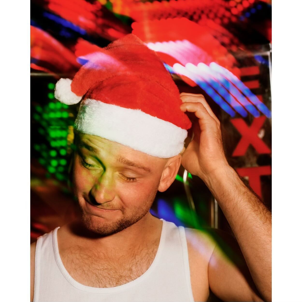 Close-up portrait of a smiling adult male wearing a Santa hat in front of a colorful, blurred background with neon lights. 