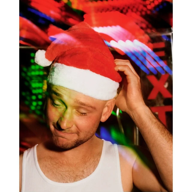 Close-up portrait of a smiling adult male wearing a Santa hat in front of a colorful, blurred background with neon lights. 