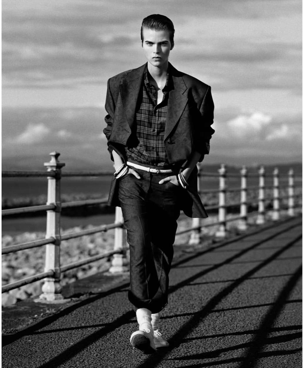 Monochrome full shot of a slender adult with hands in pockets walking confidently along a paved path by a pier, wearing smart casual attire.