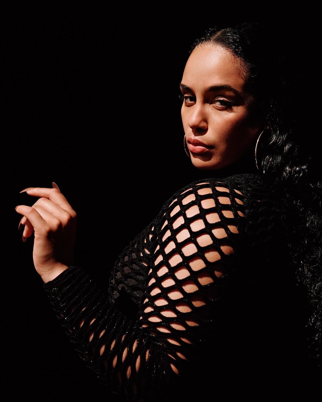 Portrait of an adult female model in a black fishnet top with hoop earrings, posed against a dark background in studio lighting.
