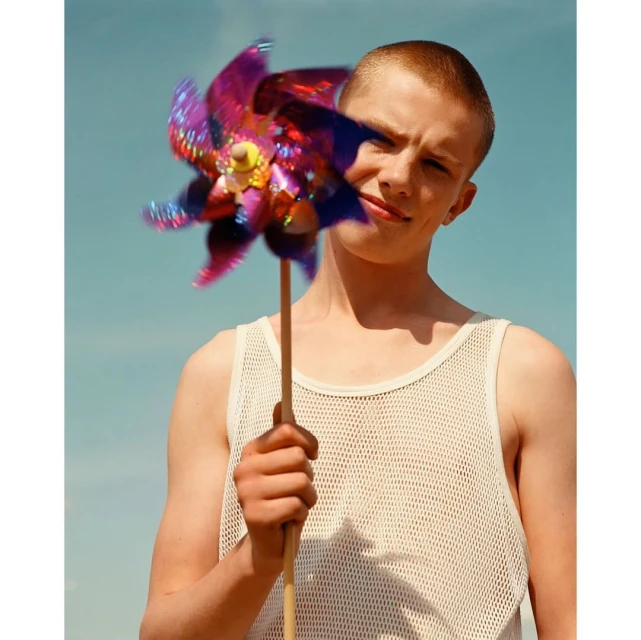 Young adult holds colorful pinwheel outdoors on sunny day. Head and shoulders shot, clear blue sky and sandy ground. Mesh tank top.