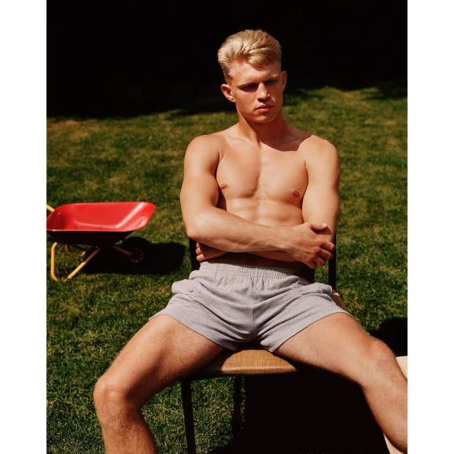 Shirtless man with blond hair sits on a chair in a grassy outdoor setting with a red wheelbarrow visible in the background.