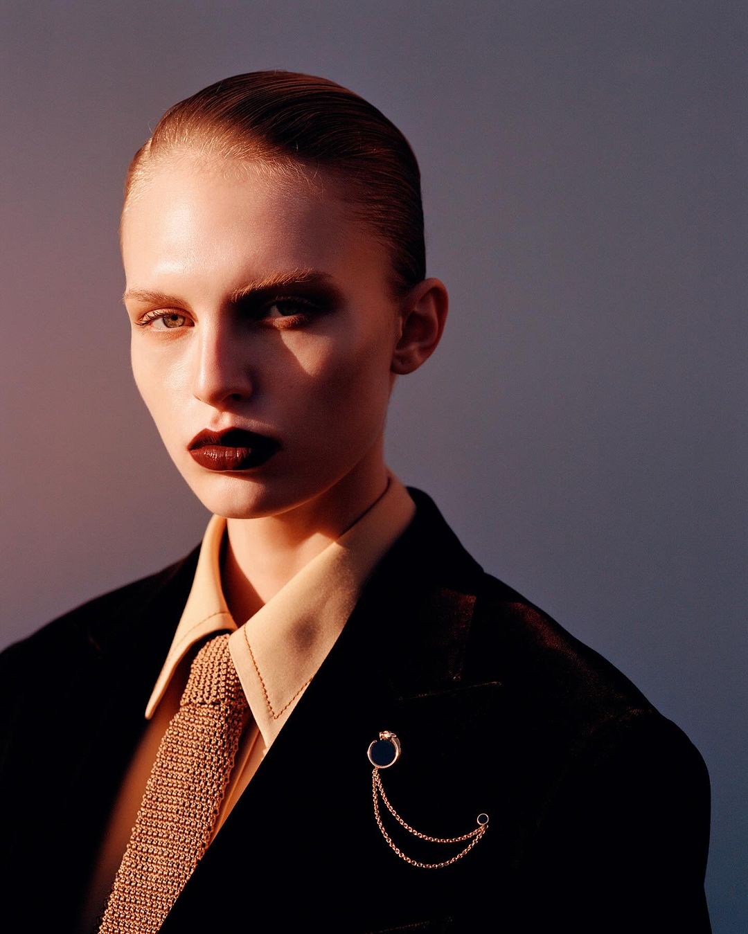 Close-up of a stylish adult female in a black suit and tie, with dark lipstick and slicked-back hair, posing against a muted background.