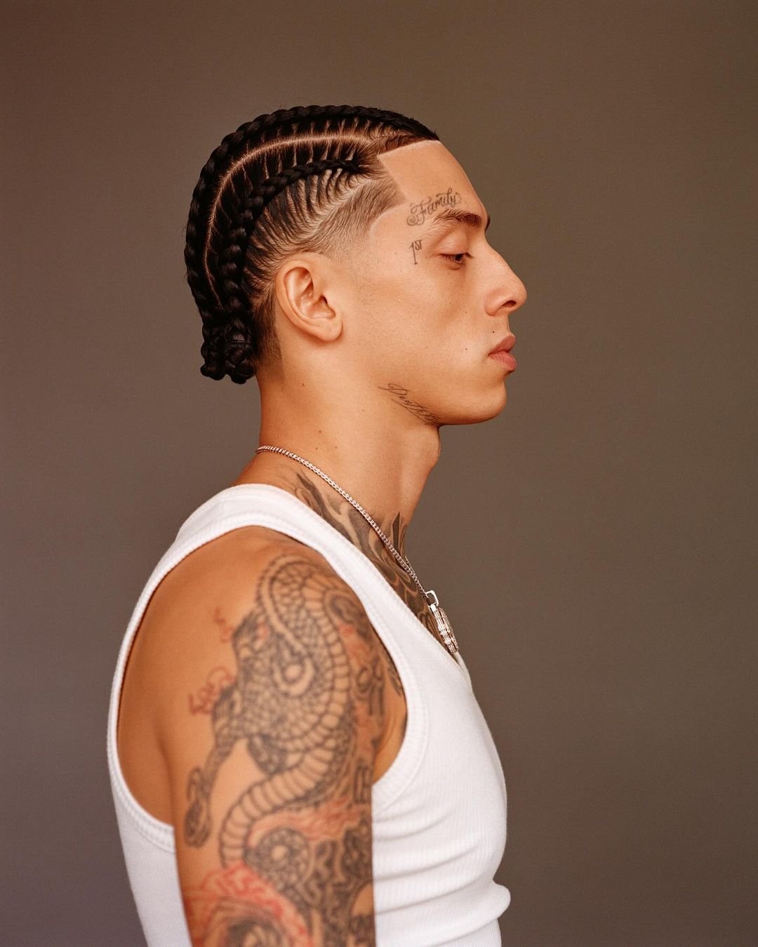 Profile portrait of a young adult male with braided hair, tattoos on his face and arm, wearing a white tank top against a neutral backdrop.