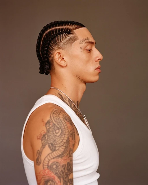 Profile portrait of a young adult male with braided hair, tattoos on his face and arm, wearing a white tank top against a neutral backdrop.