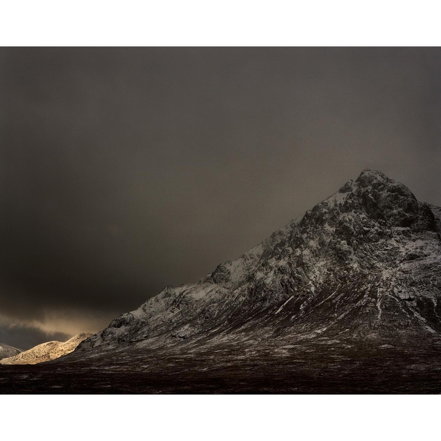 Snow-dusted mountain slope under a heavy, dark overcast sky with a patch of low evening light on the horizon.