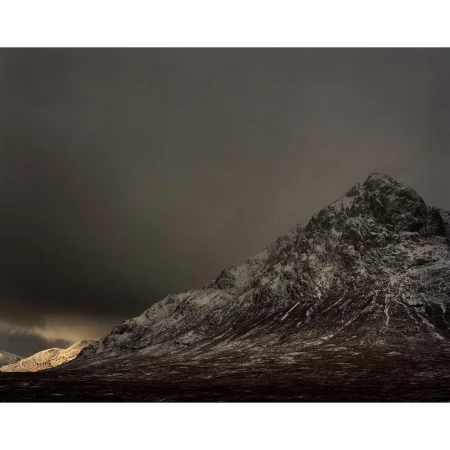 Snow-dusted mountain slope under a heavy, dark overcast sky with a patch of low evening light on the horizon.