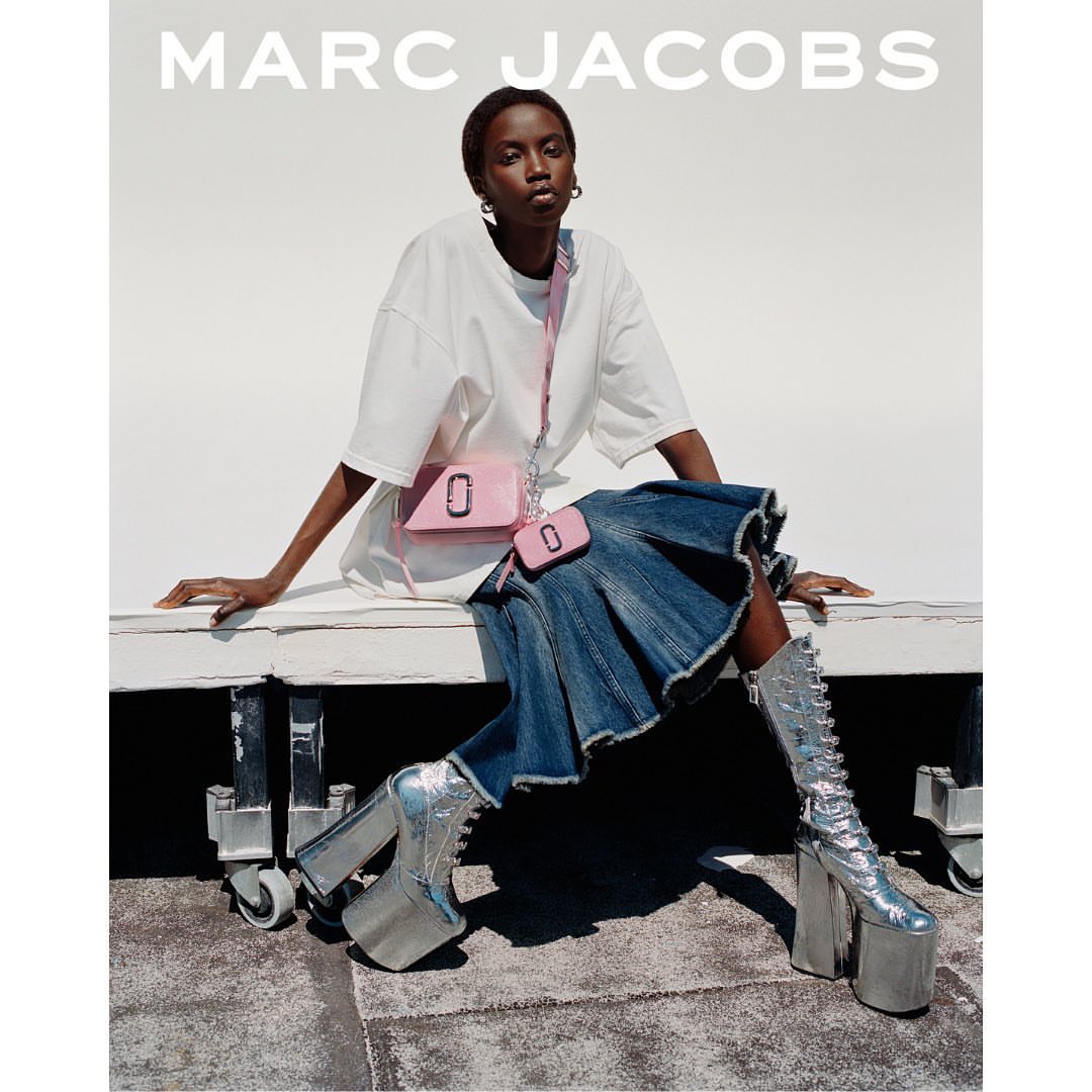 Fashion shot of a woman in platform boots sitting on a white surface wearing a t-shirt, denim skirt, and pink purse by Marc Jacobs.
