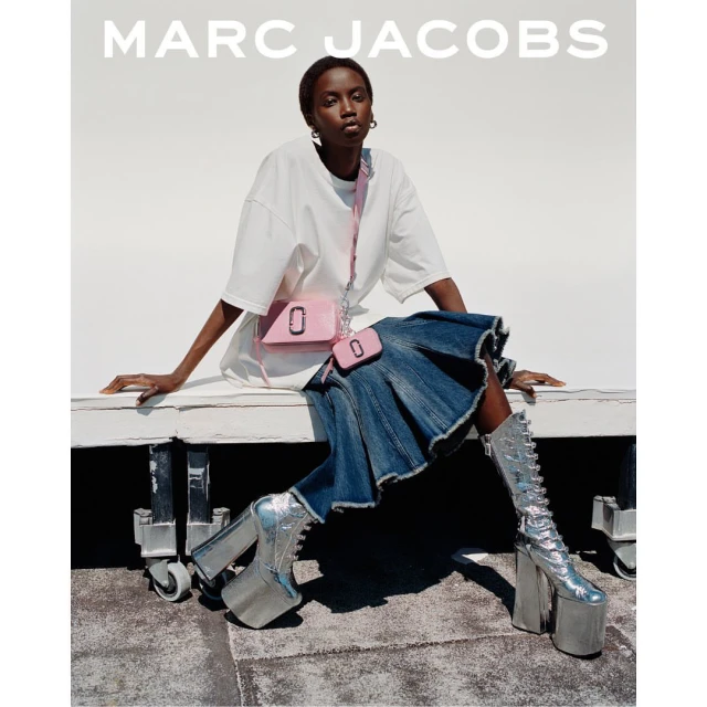 Fashion shot of a woman in platform boots sitting on a white surface wearing a t-shirt, denim skirt, and pink purse by Marc Jacobs.