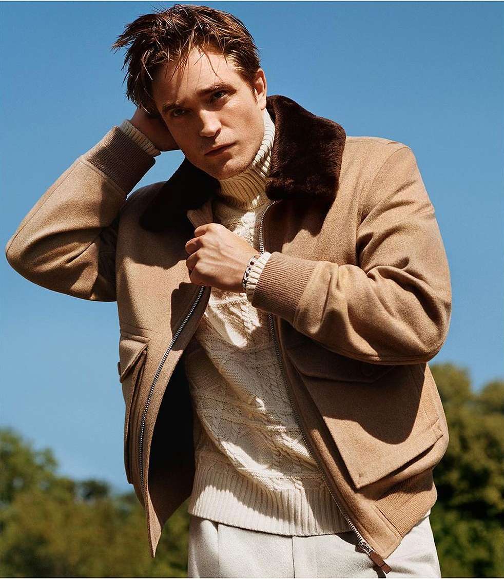 Robert Pattinson wearing a tan bomber jacket with brown collar and cable-knit sweater under a clear blue sky.
