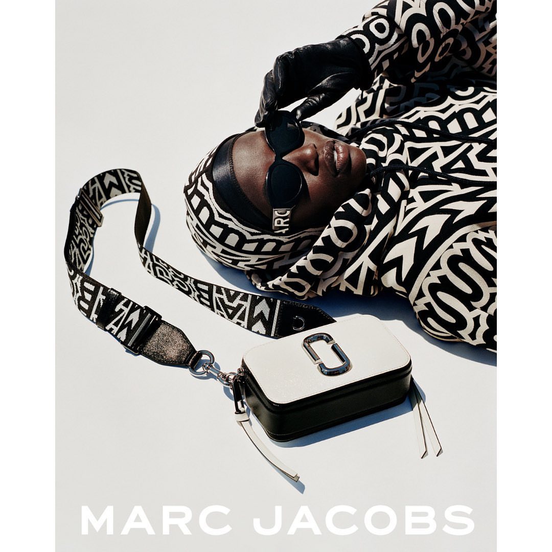 Model wearing black and white patterned hoodie and sunglasses with a white and black camera bag on a white background