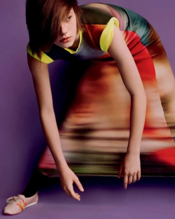 A model with short reddish-brown hair wearing a colourful patterned dress and sneakers poses against a plain purple background.