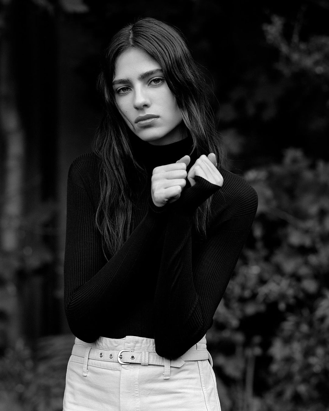 Black and white portrait of a woman with long hair wearing a black turtleneck and beige pants, set against a blurred outdoor backdrop.