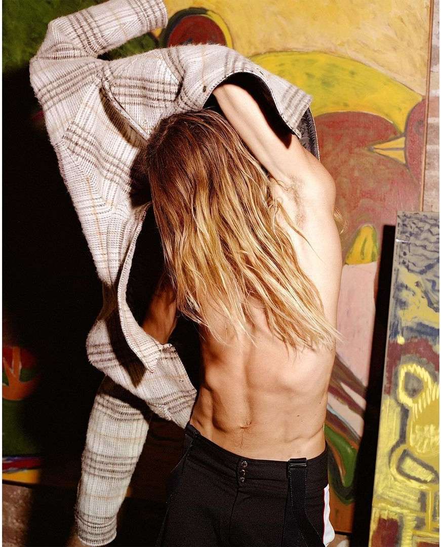 Shirtless man with long blonde hair puts on a plaid blazer indoors against a background of colorful modern art paintings.