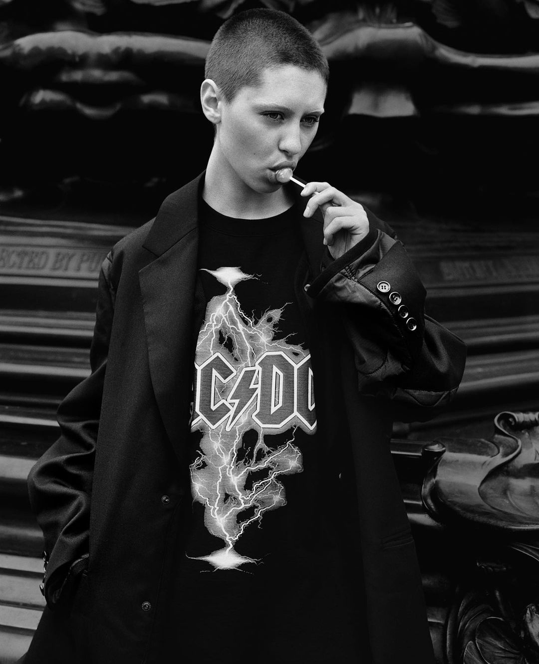 Teen wearing a black AC/DC T-shirt and oversized blazer poses outdoors with a lollipop. Fashion editorial shot in black and white.