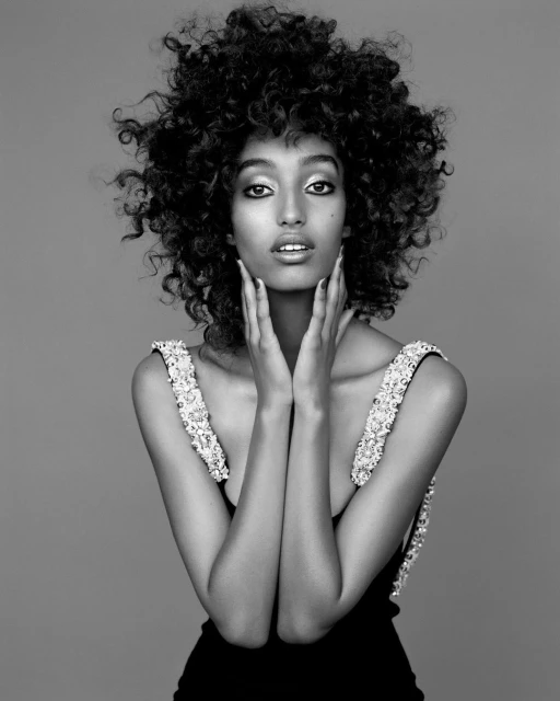 Striking black-and-white portrait of a dark-skinned woman with large curly hair, wearing an elegant black dress with jeweled straps.