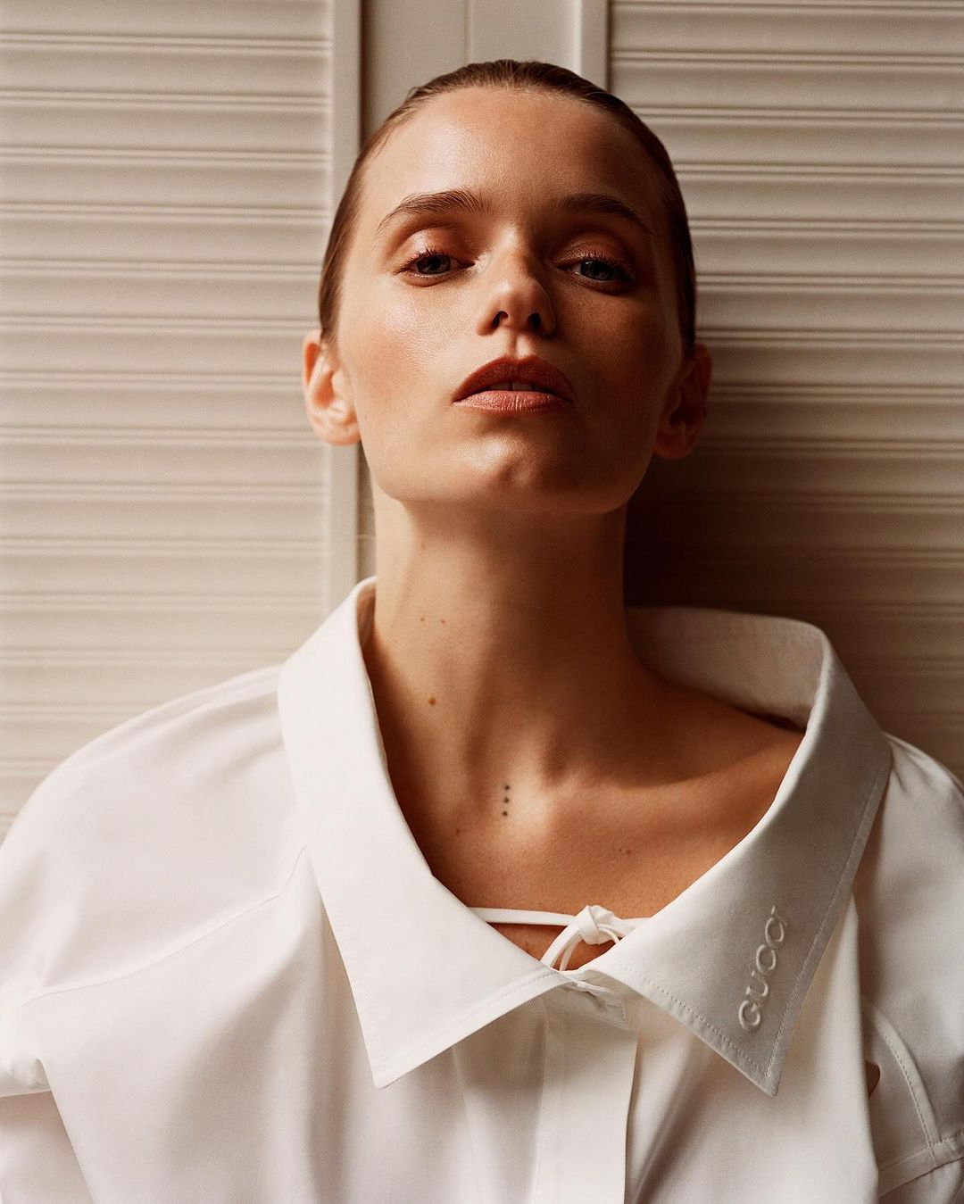 Close-up portrait of a fair-skinned woman with neutral expression, wearing a white shirt with a large collar, set against a paneled backdrop.