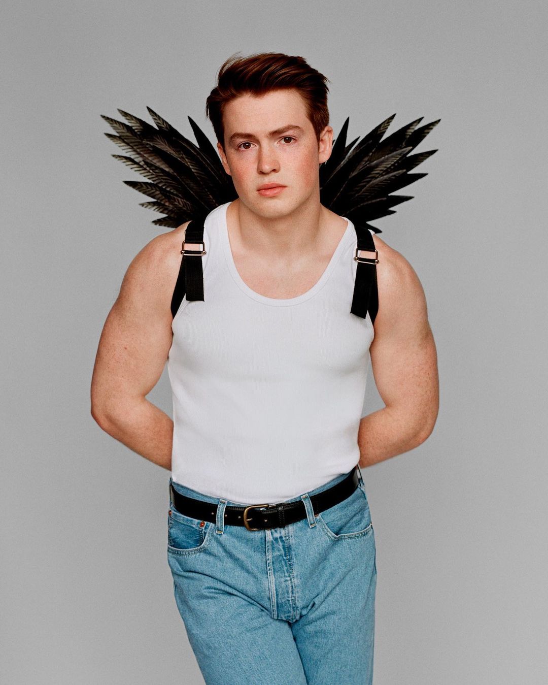 Portrait of a young man with light skin, wearing jeans and a white tank top, posing with dark feather wings against a grey background.