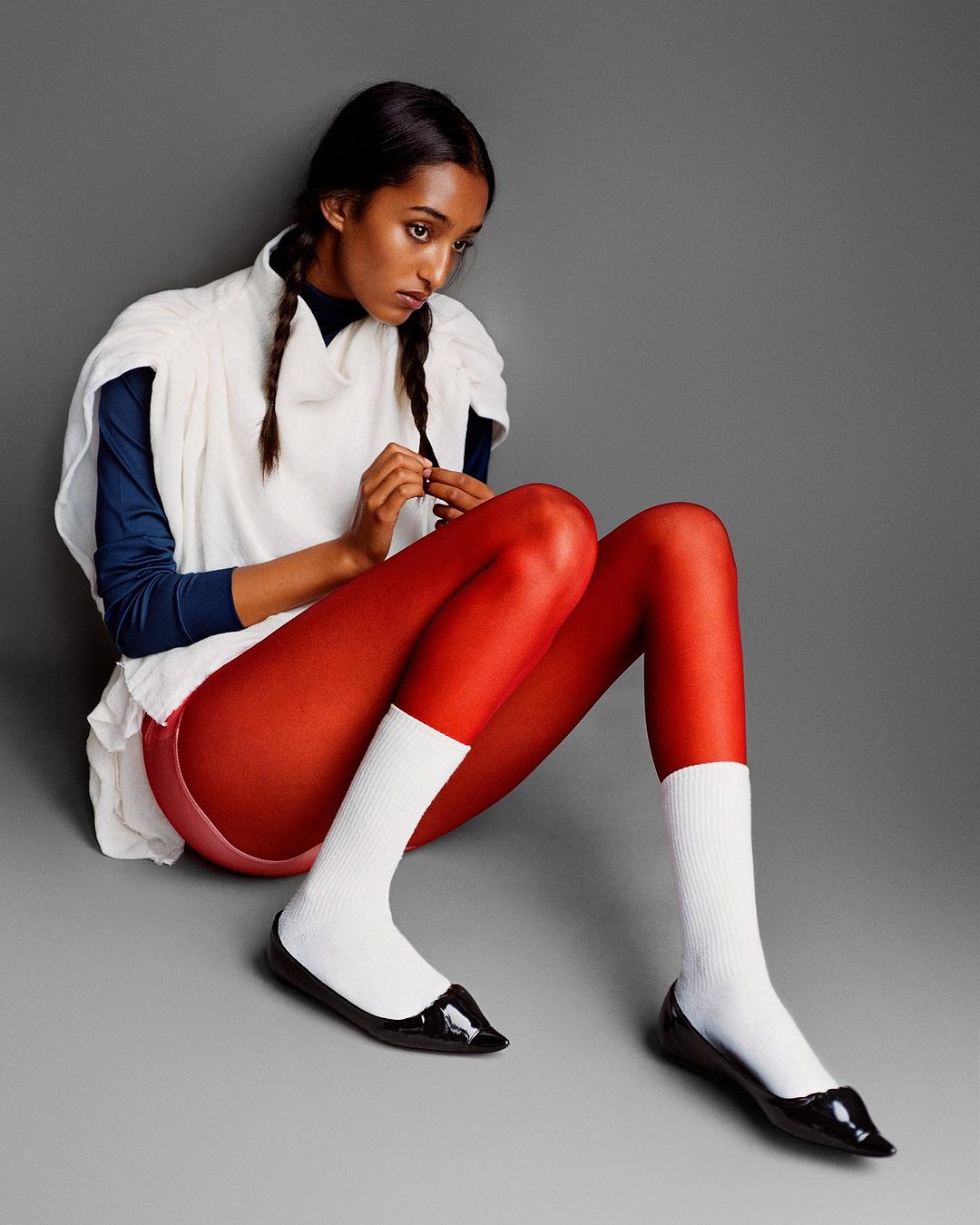 A woman with braids sits against a gray wall in red tights, white socks, and black shoes, wearing a white top and navy blue long-sleeve shirt.