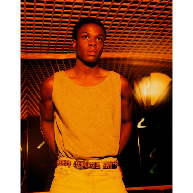 An African-American man in a tank top poses indoors with warm orange lighting. He wears light pants and a patterned belt, looking upward.