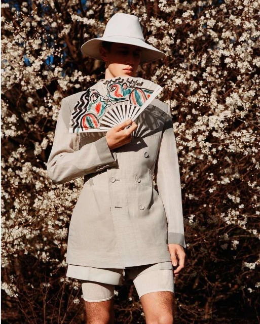A slender person in vintage-inspired fashion, wearing a hat and jacket, holding a hand fan, standing outside among flowering trees.